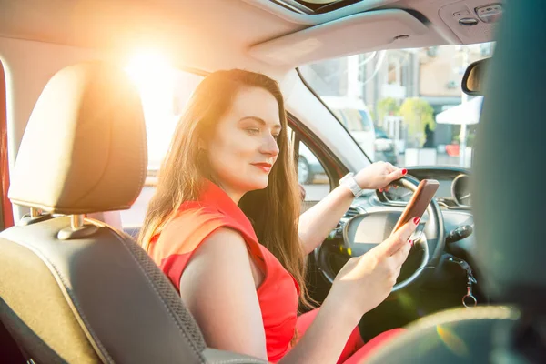 バックビュー運転中にタッチスクリーンのスマートフォンやハンドルを握って車の中で白人の若い女性ドライバー。日没バックライト。選択的フォーカス、コピースペース。景色の中. — ストック写真