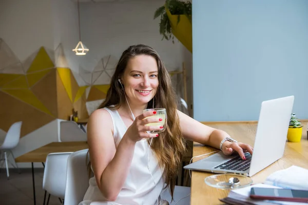 Retrato de una joven mujer de negocios sonriente con café haciendo una videollamada usando laptop y auriculares sentados en la cafetería, coworking. Trabajo remoto, redes sociales, blogs. Concepto de teletrabajo. Autónomo . —  Fotos de Stock