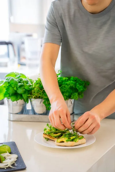 Close up man hands making a healthy meal of avocado toast and vegetables putting just picked greenery from homegrown garden on kitchen. Breakfast idea. Vegetarian and vegan diet. Vertical. Copy space. — Stock Photo, Image