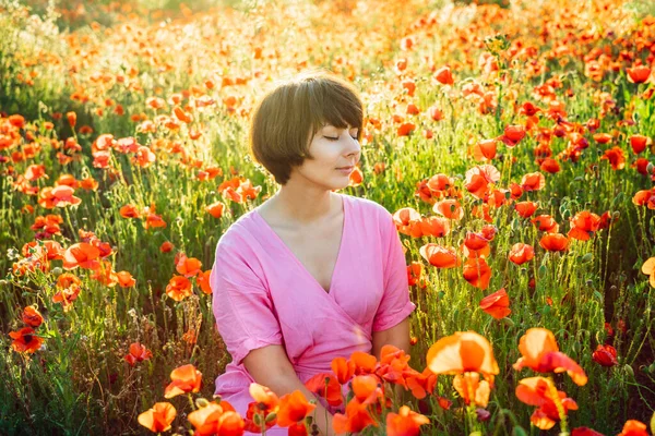 Uma jovem mulher feliz em um vestido rosa com olhos fechados, relaxando em papoilas vermelhas flores prado em luz do pôr do sol. Um simples prazer para a saúde mental. Relaxamento natural. Foco seletivo. Espaço de cópia . — Fotografia de Stock