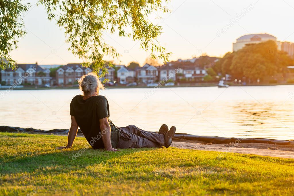 Back view gray hair mature man sitting on the green grass at the bank of park's lake and enjoying the sunset. A simple pleasure for mental health. Nature relaxation. Selective focus. Copy space