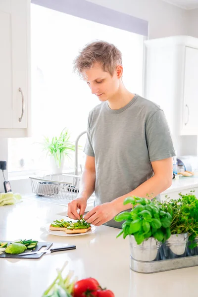 Young caucasian man making a healthy meal of avocado toast and vegetables putting just picked greenery from homegrown garden on the kitchen. Breakfast ideas. Vegetarian and vegan diet. Copy space. — Stock Photo, Image