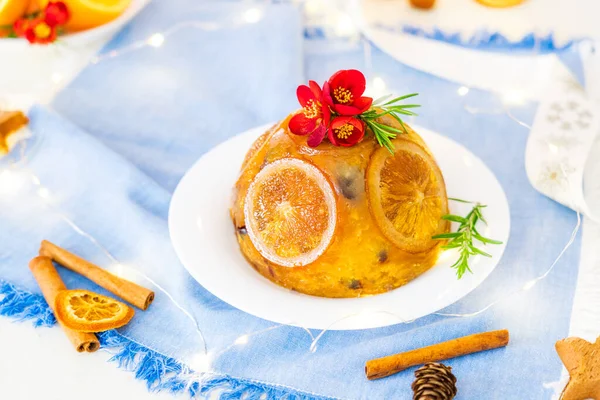 Traditional English Christmas pudding decorated with oranges and fresh flowers in a festive table setting with fruits, spices and garland lights on blue napkin background. Copy space. Magical mood. — Stock Photo, Image