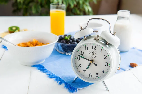 Zeit zum Frühstück. Wecker mit gesundem Essen im Hintergrund. Schüssel mit Müsli, Nüssen und Beeren, Milch, frischem Saft auf blauer Serviette und weißem Holztisch. Ernährungsplanung und gesunde Ernährung. — Stockfoto