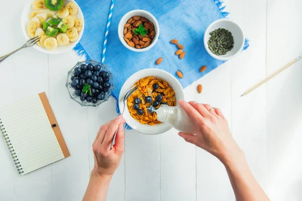 Top view female hands adding milk to cereals bowl with berries. Morning habits. Day planning and healthy eating. Time for breakfast. Alarm clock with healthy food and notebook on white wooden table. — Stock Photo, Image