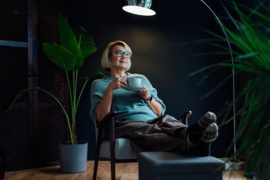 Middle aged woman relaxing with cup of hot drink in scandy style hygge interior home. Cozy comfortable winter or autumn weekends. Take a break and relax. Lady enjoy calm mood, well being alone