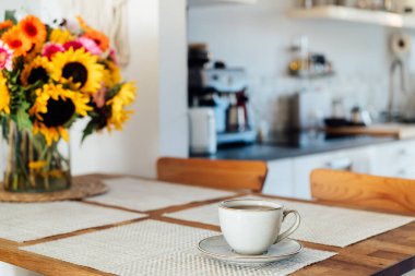 Bir fincan kahve, çay ve vazoya odaklanın. Mutfağın samimi, modern iç kısmında ahşap masa üzerinde büyük ayçiçekleri buketi olsun. Samimi Tasarım ev dekorasyonu. Yaz havası, günaydın rutini. Seçici odak