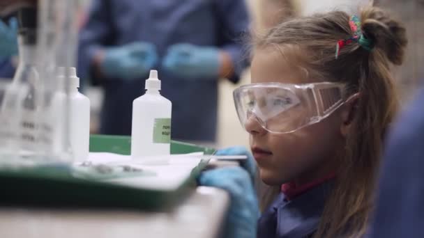 Laboratorio, un scoolgirl en gafas de seguridad examina un líquido en un frasco de vidrio, experiencia química, Educación de los niños. — Vídeos de Stock