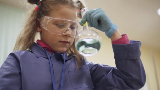 Laboratorium, een scoolgirl in beschermende bril onderzoekt een vloeistof in een maatkolf van glas, chemische ervaring, onderwijs kinderen. — Stockvideo