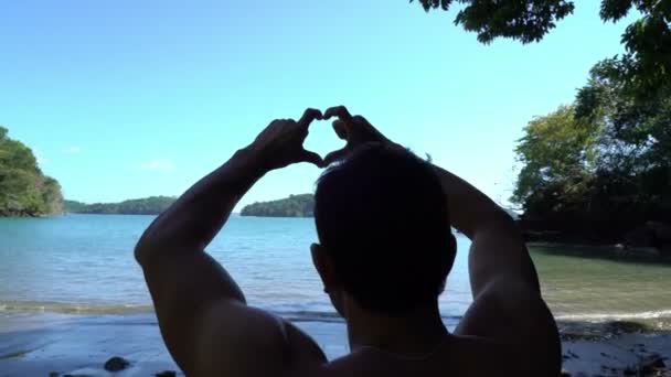 Joyeux jeune homme assis sur la plage sauvage magnifique faisant cadre avec les mains. Portrait d'un homme regardant à travers une caméra imaginaire faite avec des doigts. Les gens s'inspirent du paysage, du travail de photographe — Video