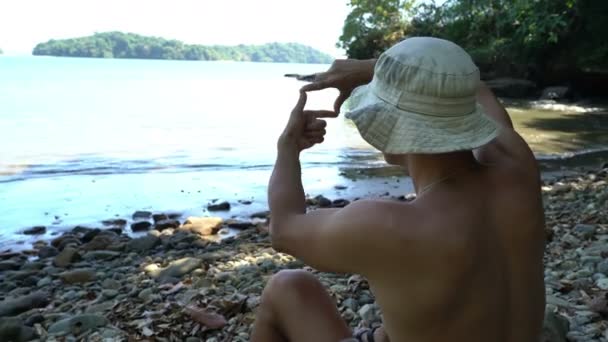 Веселый молодой человек, сидящий на диком красивом пляже, делая раму руками. Портрет мужчины, смотрящего через воображаемую камеру, сделанную пальцами. Вдохновение от пейзажа, работы фотографа — стоковое видео