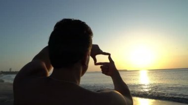 Fotoğraf çerçevesi şaşırtıcı, elleri ile sahilde günbatımı yapma kıvırcık saçlı Genç adam