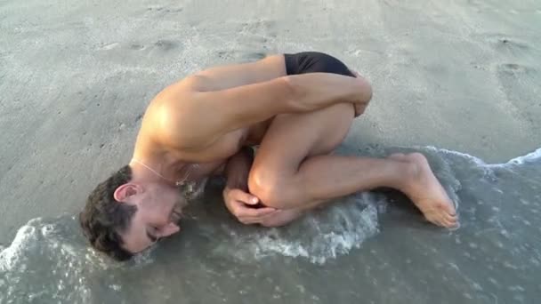 Чоловік лежить на пляжі біля води в позі ембріона. Концепція еволюції. Народження. Походження від — стокове відео