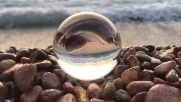 Благодатний фоновий пейзаж всередині кришталевого м'яча лежить на пляжних каменях, заході сонця та океанічних хвилях всередині — стокове відео