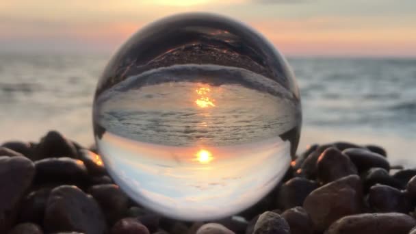 Mooie achtergrond landschap binnen kristallen bol lag op strand stenen, zonsondergang en oceaan golven binnen — Stockvideo