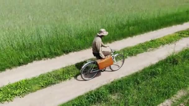 Человек едет на велосипеде по проселочной дороге в зеленом поле, держа чемодан в руке, смешное путешествие — стоковое видео