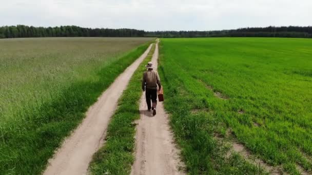 Muž sám kráčí po venkovské silnici v zeleném poli, drží kufr v ruce, legrační cestovní koncept — Stock video