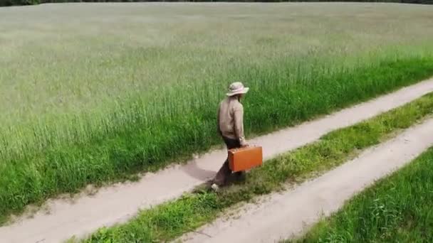 Hombre solo caminando por el camino del campo en un campo verde, sosteniendo la maleta en una mano, concepto de viaje divertido — Vídeo de stock