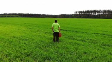 Kafası karışmış üzgün bir adam yeşil bir tarlada duruyor, elinde bavulla, İHA 'dan havadan ateş ediyor.