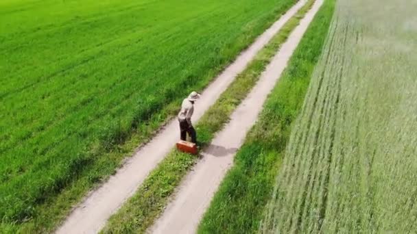 Kırsal yolda tek başına yürüyen bir adam dinlenmek için yeşil bir arazide durur ve bavuluna oturur. — Stok video