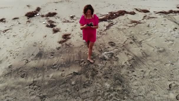 Vrouw in rode jurk drone piloot lopen in het water door zand strand aan de kust bij zonsondergang — Stockvideo