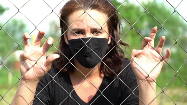 Metal tel örgülerin arkasında tıbbi koruyucu maskeli bir kadın var. Sosyal mesafe kavramı ve...