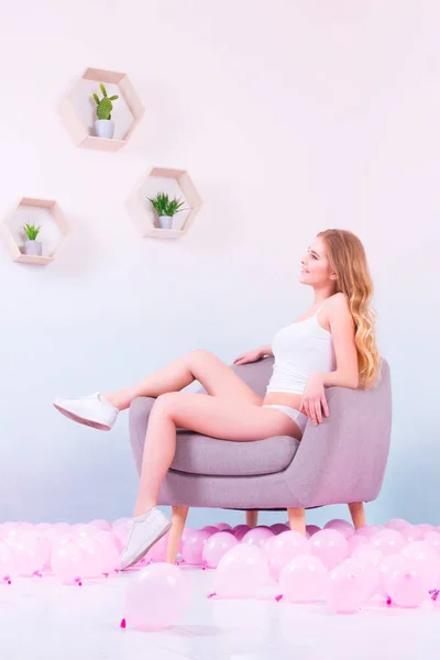 Sporty girl in white underwear and white sneakers sitting on the purple armchair, smiling and looking at the plants on the wooden shelves in her room with pink balloons on the floor