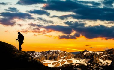 Kaya tırmanışı ekipmanlarıyla tırmanan bir adam yüksek kayalıklarda tırmanıp bir sonraki adımı atıyor kayalık tepeye ulaşıyor. Yan görüş. Dağların panoramik manzarası günbatımı ve bulutlar ile gökyüzü.