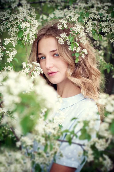 Güzel mutlu genç kadın çiçekli bir bahar bahçesinde koku zevk. Mavi gözlü ve mavi elbiseli sarışın.