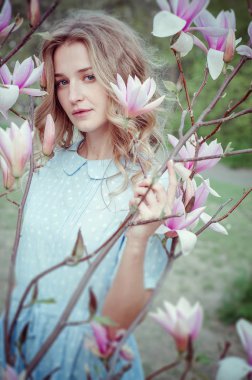 Güzel mutlu genç kadın çiçekli bir bahar bahçesinde koku zevk. Bereketli ağaç manolyaları, büyük çiçekler. Mavi gözlü ve mavi elbiseli sarışın.