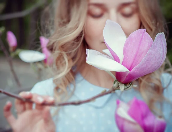 Güzel mutlu genç kadın çiçekli bir bahar bahçesinde koku zevk. Bereketli ağaç manolyaları, büyük çiçekler. Mavi gözlü ve mavi elbiseli sarışın.
