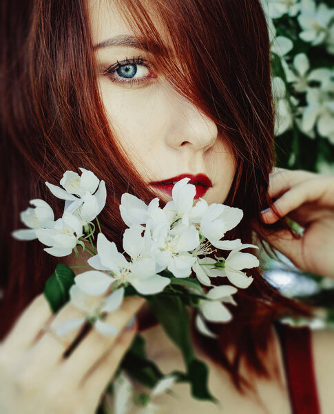 Beautiful young woman enjoying smell in a flowering spring garden.Beutiful tree , flowers. Red hair, blue eyes and red dress,holds the flowers in their hands.
