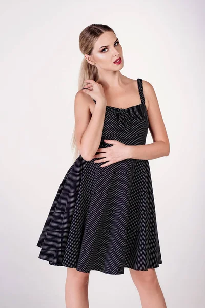 Açık saçlı genç çekici seksi sarışın kadın model, topuklu kısa siyah elbise ve sandalet giyen, poz ve beyaz stüdyoda dans