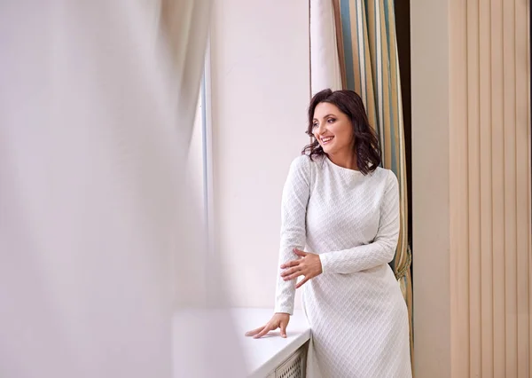 Beyaz elbise giyen koyu saçlı güzel orta eged gülümseyen kadın model, ışık odasında mavi kanepe üzerinde poz.