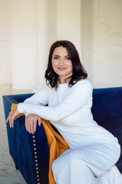 Beyaz elbise giyen koyu saçlı güzel orta eged gülümseyen kadın model, ışık odasında mavi kanepe üzerinde poz.