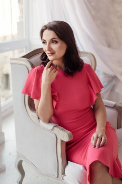 Pembe elbise giyen koyu saçlı güzel orta eged gülümseyen kadın modeli, klasik odada pencere yakınında poz.