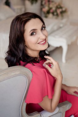 Pembe elbise giyen koyu saçlı güzel orta eged gülümseyen kadın modeli, klasik odada pencere yakınında poz.