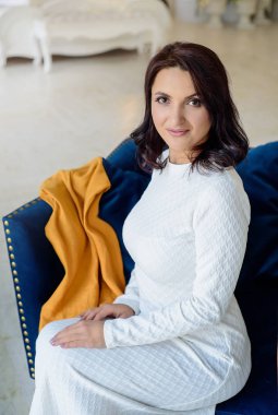 Beyaz elbise giyen koyu saçlı güzel orta eged gülümseyen kadın model, ışık odasında mavi kanepe üzerinde poz.