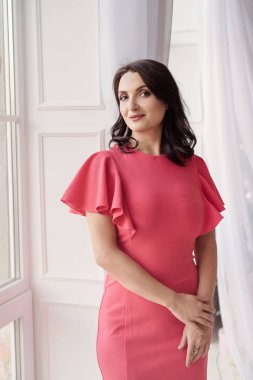 Pembe elbise giyen koyu saçlı güzel orta eged gülümseyen kadın modeli, klasik odada pencere yakınında poz.