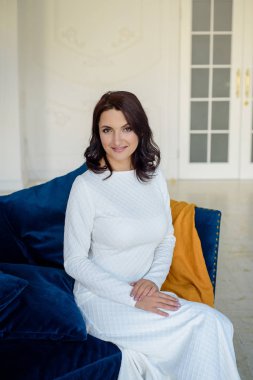Beyaz elbise giyen koyu saçlı güzel orta eged gülümseyen kadın model, ışık odasında mavi kanepe üzerinde poz.