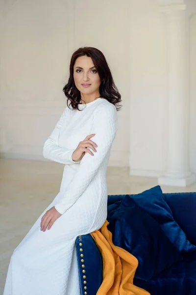 Beyaz elbise giyen koyu saçlı güzel orta eged gülümseyen kadın model, ışık odasında mavi kanepe üzerinde poz.