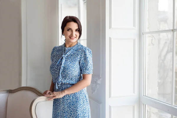 Mavi pin-up elbise giyen koyu saçlı güzel orta eged gülümseyen kadın modeli, klasik odada pencere yakınında poz.