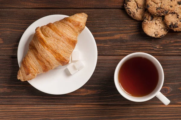 Koyu ahşap arka planda kruvasan ve kurabiyeli çay bardağı.