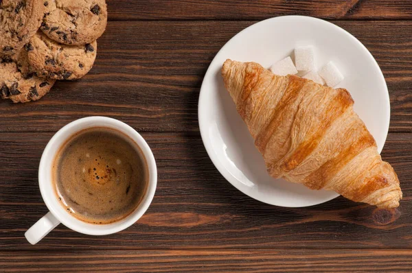 Koyu ahşap arka planda kruvasan ve kurabiyeli kahve bardağı.