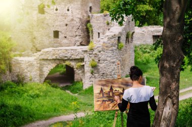 Student artist paints painting paint and brush. The picture depicts an old fortress in the city of Kamyanets-Podilsky. clipart