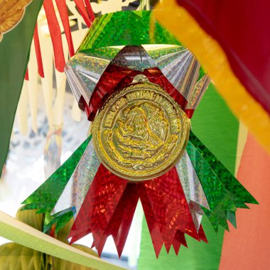 Yeşil kurdeleli altın ve kırmızı bir Meksika bayrağı. Kurdele bir fiyonkta bağlı. Bayrak duvarda asılı.