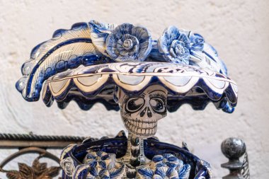 A blue and white ceramic skull with flowers on top of it. The skull is placed on a table and the flowers are arranged around it. Scene is eerie and somewhat macabre. City of Puebla, Mexico, crafts