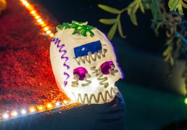 A skull with purple and blue decorations is sitting on a table. The skull is decorated with purple and blue ribbons. Day of the Dead celebration in Mexico, papel picado crafts, offerings, La Catrina