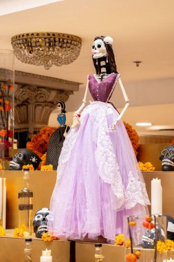 A skeleton is wearing a beautiful purple dress and standing in front of a table filled with various skulls. Day of the Dead celebration in Mexico, papel picado crafts, offerings, La Catrina