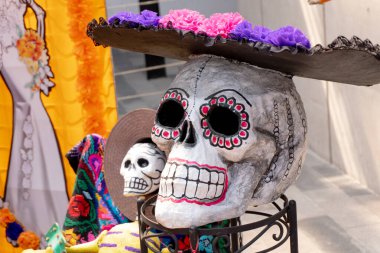 A skull with a flowery hat on top of it. The skull is surrounded by other skulls and a woman's dress. Day of the Dead celebration in Mexico, papel picado crafts, offerings, La Catrina